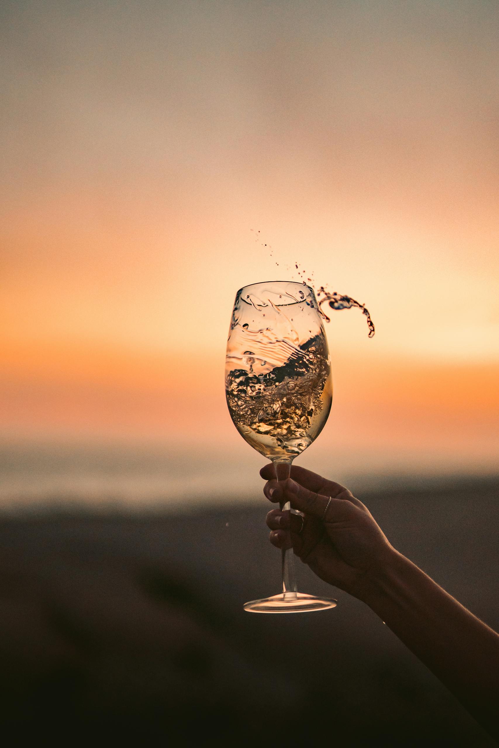 Hand holding a splashing wine glass against a vibrant sunset beach backdrop.